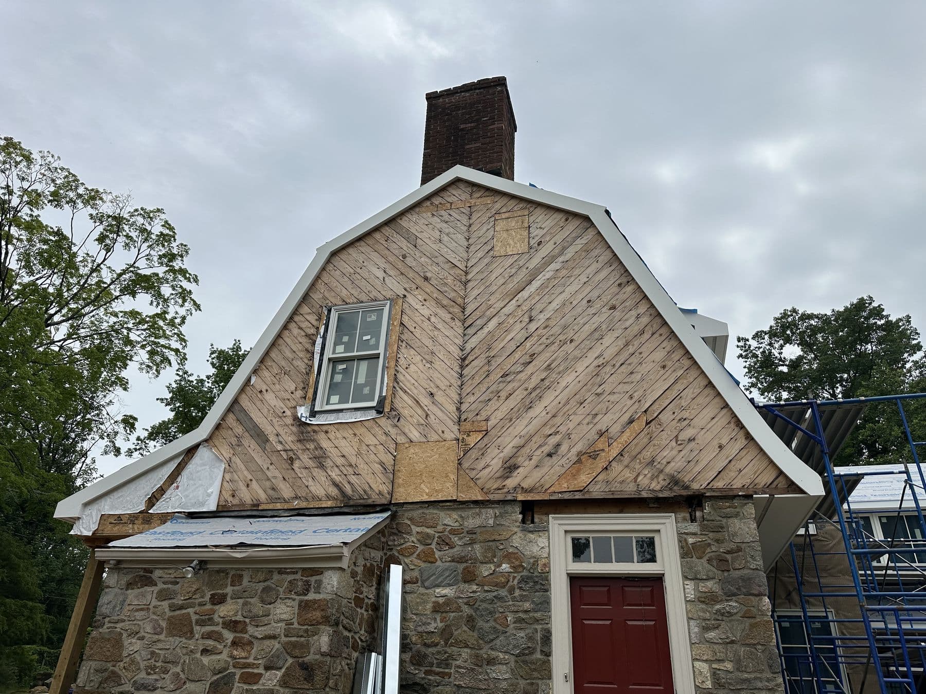 Gambrel end wall of the stone house with old wood boards exposed and a red front door below