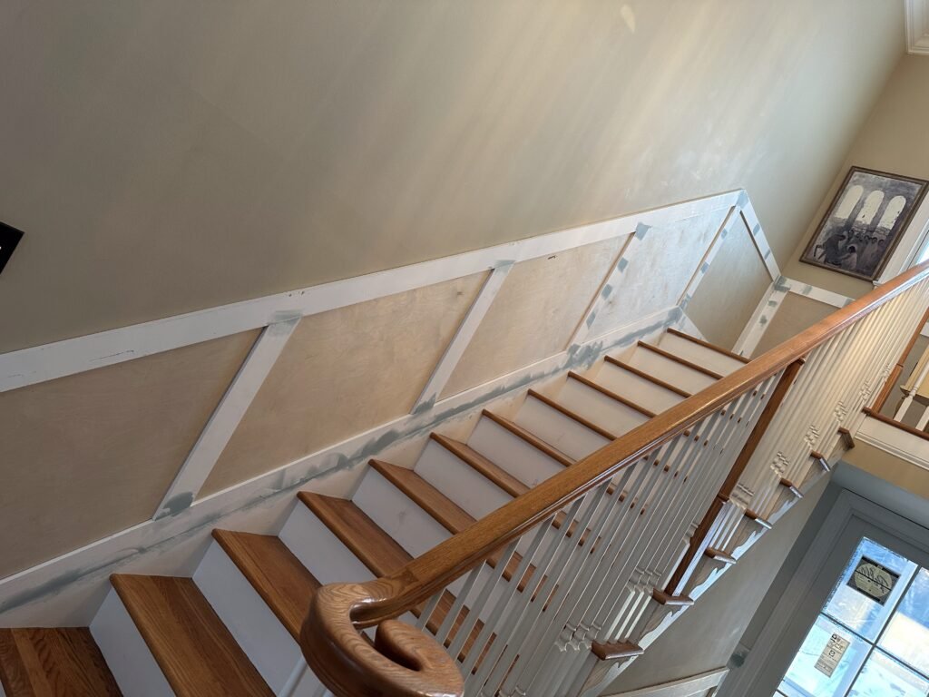 Oak staircase with wood handrail and balusters beside paneled wainscoting under construction