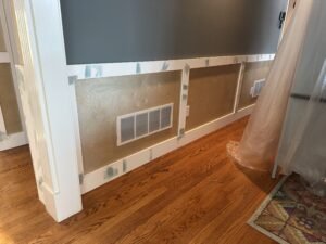 Board-and-batten wainscoting under construction with plywood panels and white rails below a gray wall
