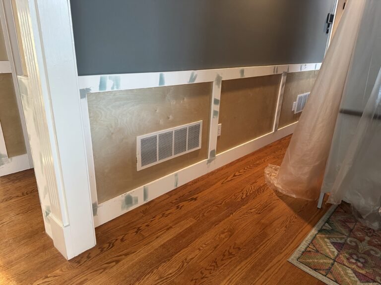 Board-and-batten wainscoting under construction with plywood panels and white rails below a gray wall