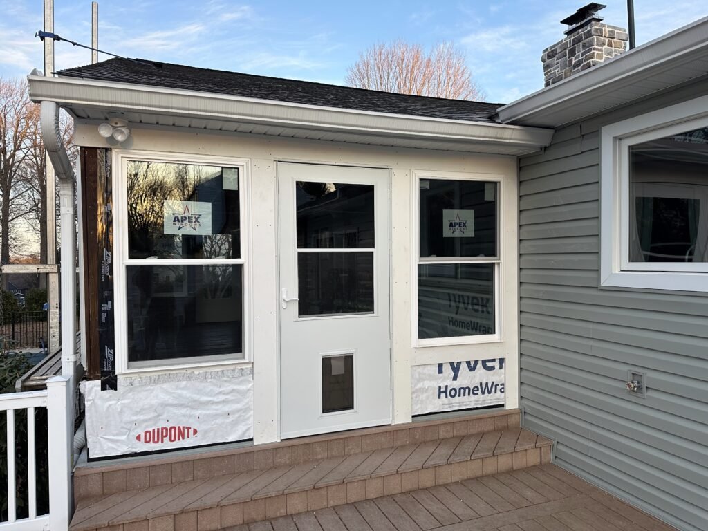 Sunroom porch addition under construction with a white door, gray siding, and a composite deck