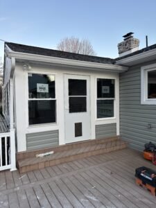 Enclosed sunroom porch addition with a white door and windows over a composite deck beside gray siding