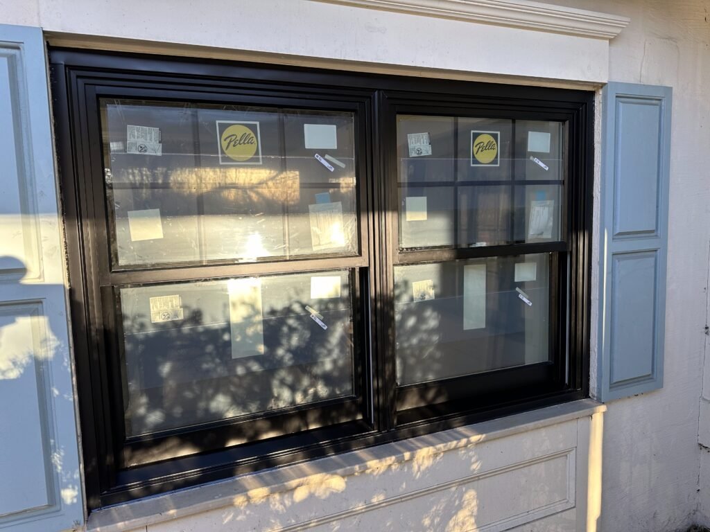 Paired black Pella double-hung windows with a decorative cornice on a cream stucco wall with sage shutters