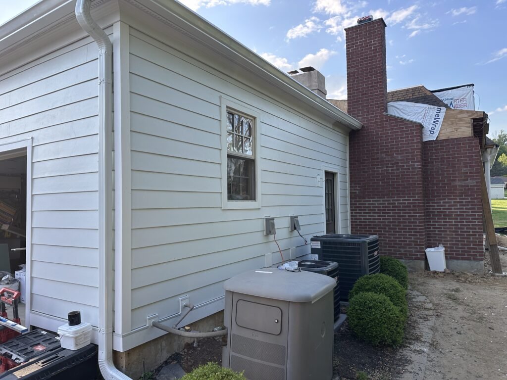 Cream fiber cement lap siding with white gutter and downspout beside a brick chimney and AC units