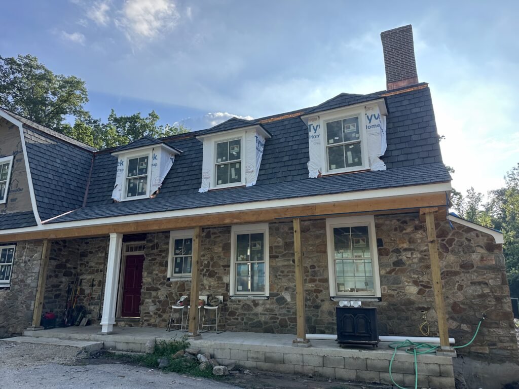 Historic fieldstone gambrel farmhouse under renovation with three dormers, a slate roof, and a porch