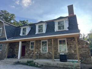 Historic fieldstone gambrel farmhouse under renovation with three dormers, a slate roof, and a porch