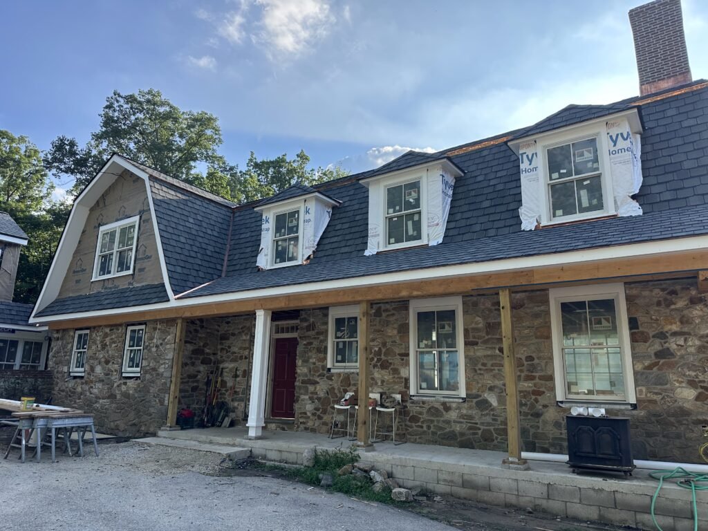 Historic fieldstone gambrel home with slate roof and dormers under renovation with a red entry door