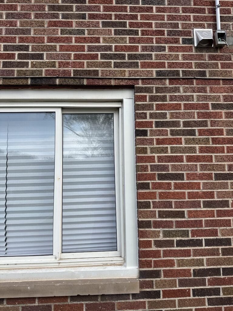 Old white sliding window and dryer vent on a red brick home before window replacement