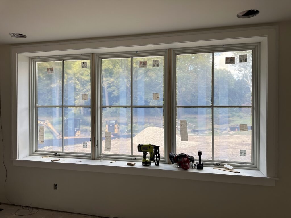 Large three-unit white Andersen window with built-up trim and a deep sill in a room under construction