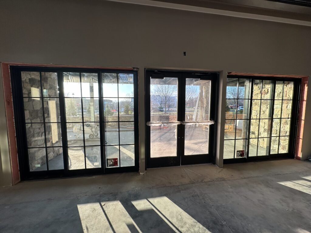 Black-framed commercial storefront windows and glass double doors seen from inside a building under renovation