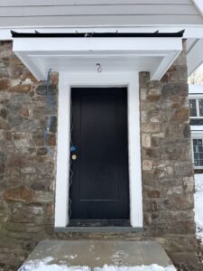 Black entry door with white trim under a portico set into a fieldstone wall with bluestone step