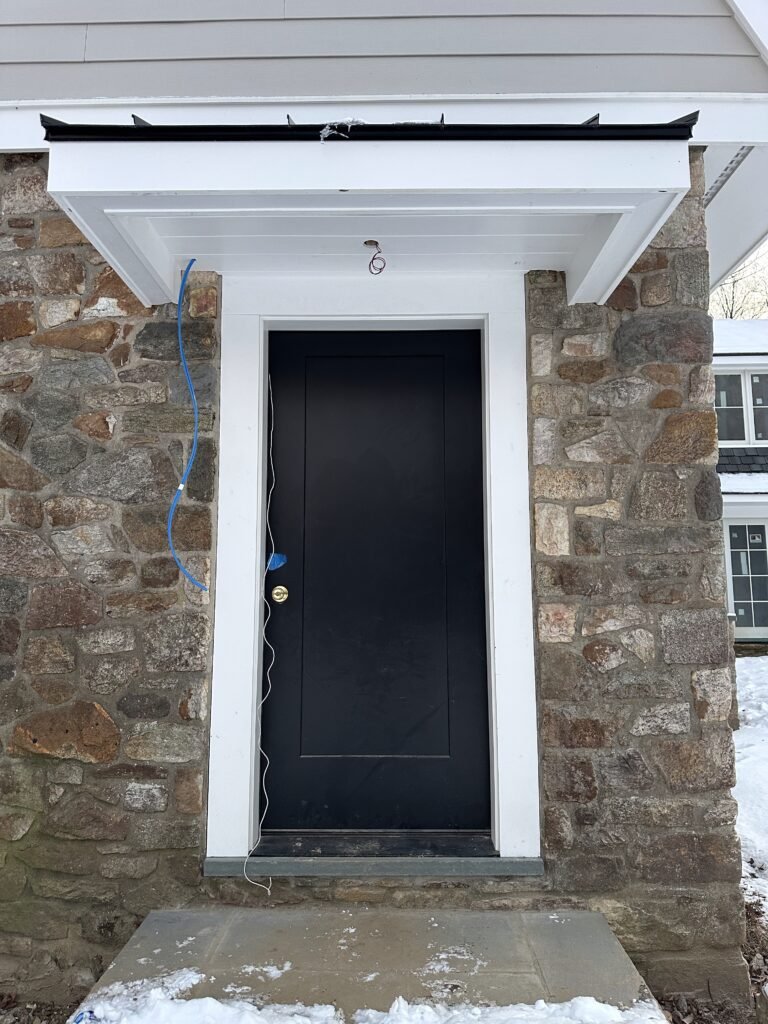 Black entry door with white trim under a portico set into a fieldstone wall with bluestone step