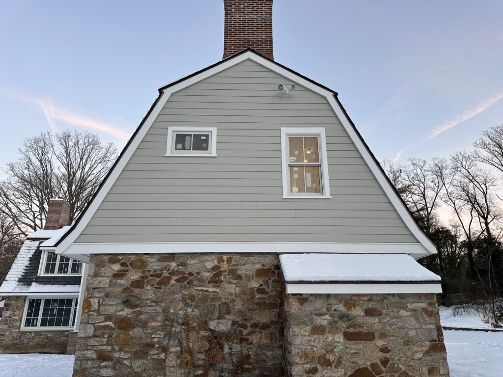 Gray clapboard gambrel gable over a fieldstone base with white-trimmed windows and a brick chimney in winter