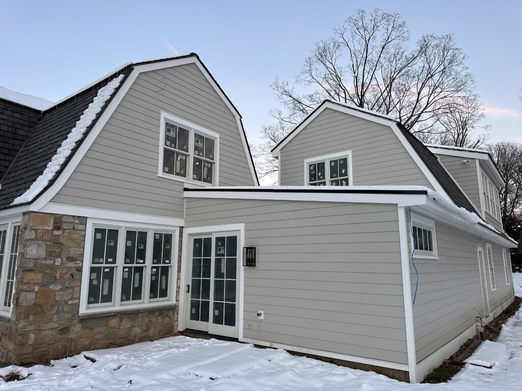 Gray gambrel-roof home with dormers, white-trimmed windows, and a stone accent in winter