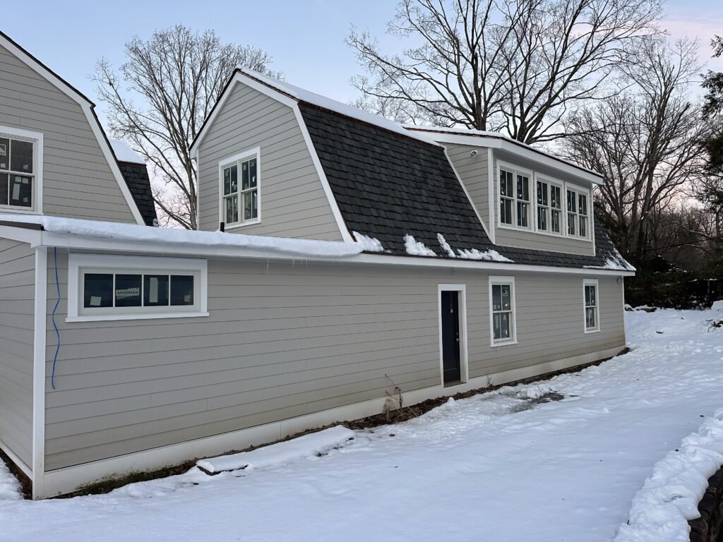 Gray clapboard gambrel-roof home with white-trimmed windows and a window dormer in winter