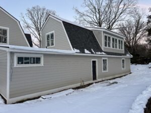 Gray clapboard gambrel-roof home with white-trimmed windows and a window dormer in winter