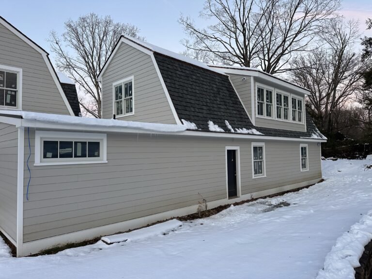 Gray clapboard gambrel-roof home with white-trimmed windows and a window dormer in winter