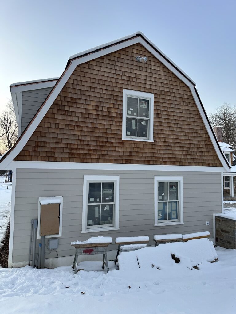 Gambrel home with cedar shake upper and gray clapboard lower siding with new white-trimmed windows in winter