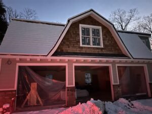 Three-bay carriage house with a cedar shake gambrel gable and garage openings under construction in winter