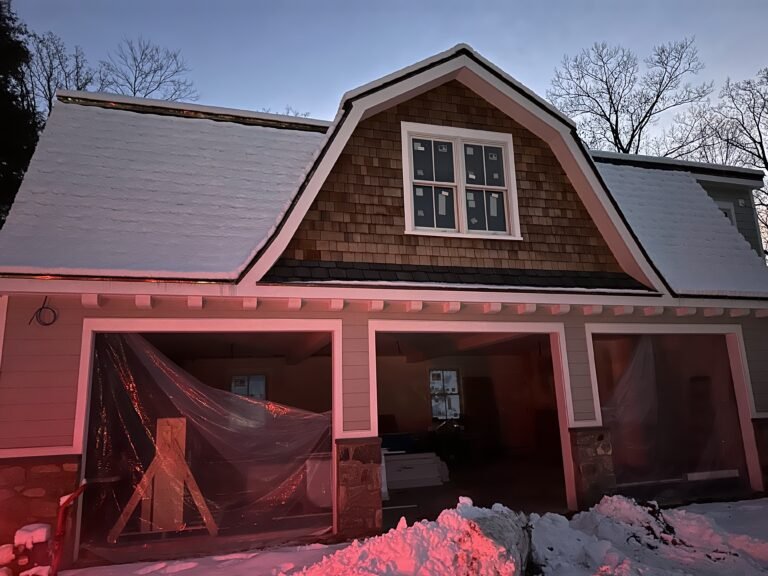 Three-bay carriage house with a cedar shake gambrel gable and garage openings under construction in winter