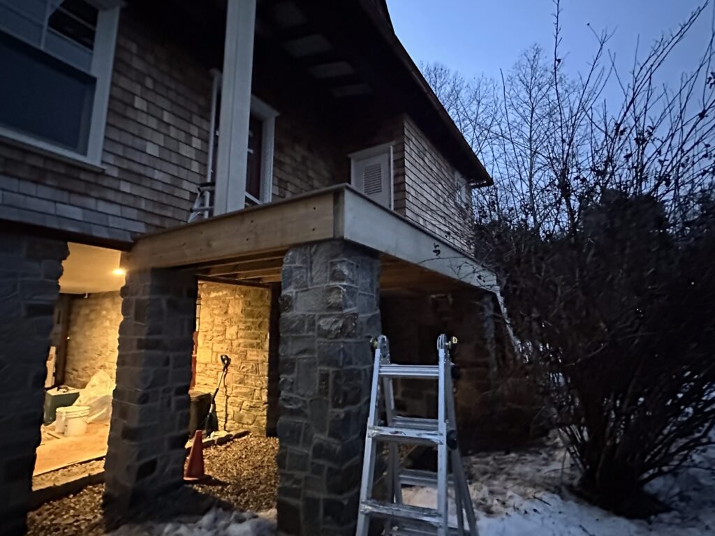 Cedar shake home with stone pier columns supporting a deck under construction at dusk