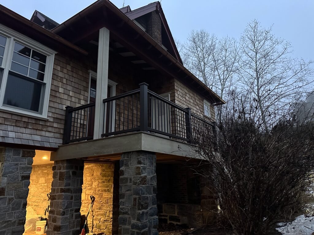 Cedar shake home with stone columns and a second-floor balcony with black metal railing at dusk