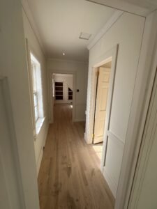 Interior hallway mid-renovation showing new white oak hardwood floors, chair rail trim, Marvin window, and bathroom tile floor visible through doorway in Downingtown PA Chester County