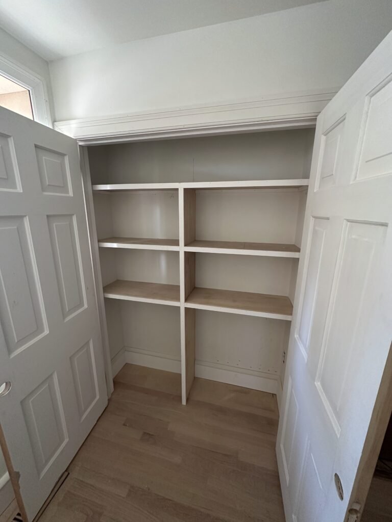 Built-in closet shelving with plywood shelves and open white bifold doors over oak flooring